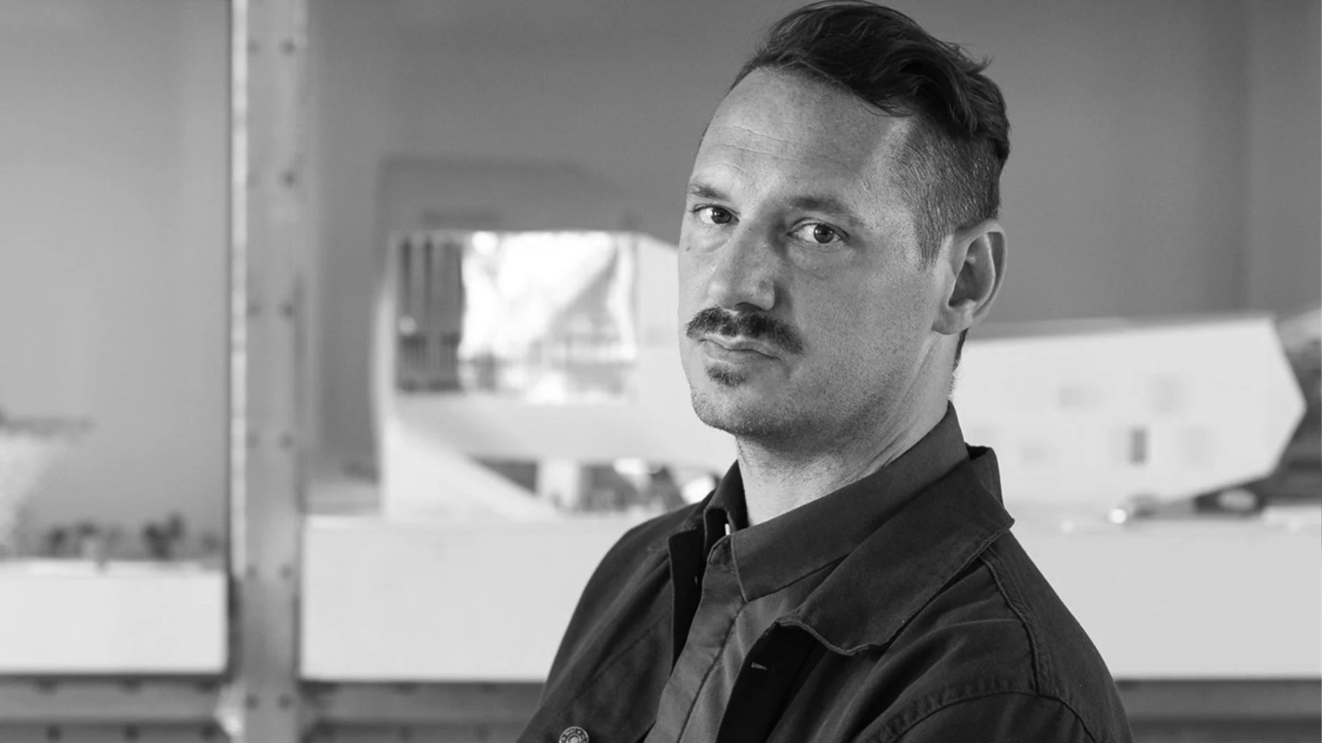 Svartvitt porträtt av en man med mustasch och mörka kläder, som tittar in i kameran.