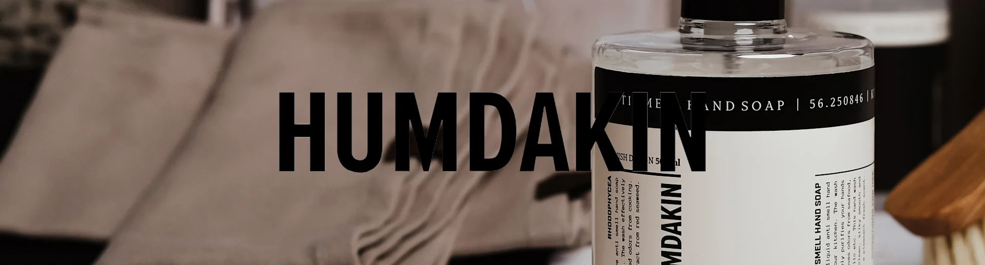 Humdakin Anti Smell Handtvål i genomskinlig flaska med svartvit etikett, mot en oskarp bakgrund med handdukar och en borste.