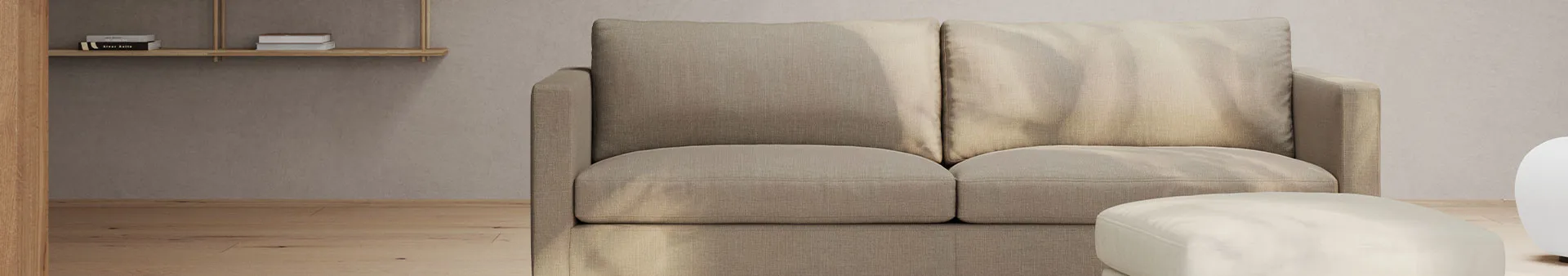 En beige tvåsitssoffa och ottoman i ett minimalistiskt rum med ljusa trägolv och en trähylla med böcker.