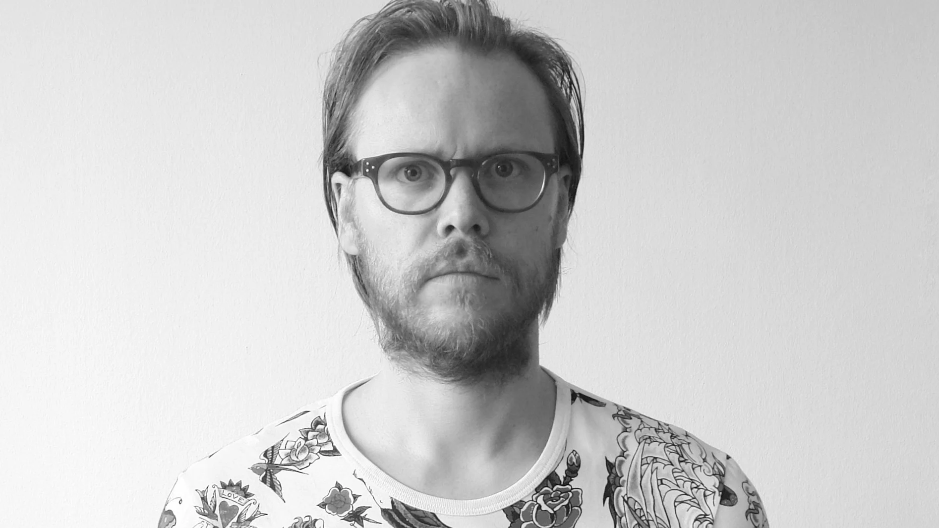 Man med glasögon och skägg i en mönstrad t-shirt, tittar rakt in i kameran.
