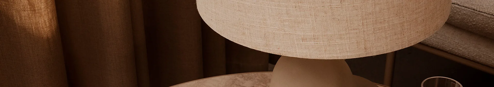 Texturerad beige lampskärm på ett bord, med mörkbruna gardiner i bakgrunden.