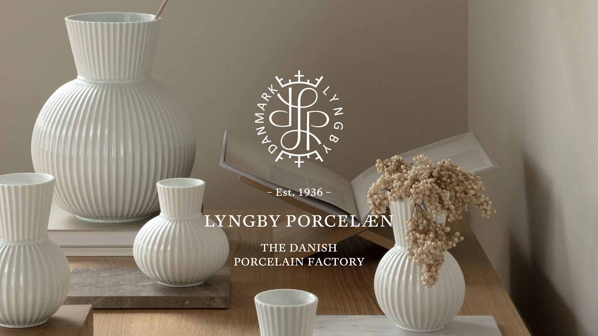 Vita räfflade porslinsvaser och koppar på träyta, en vas med torkade blommor. Lyngby Porcelæn-logotyp i bakgrunden.