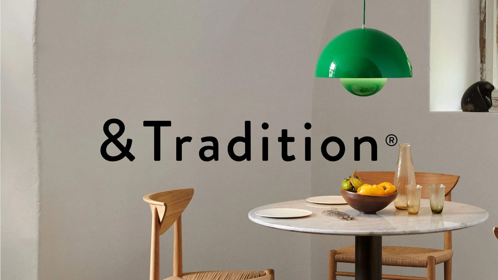 Matsal med runt marmorbord, två trästolar och en grön taklampa. Frukt i en skål, vas och glas på bordet. '&Tradition®' på väggen.