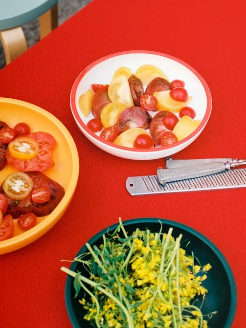 Färsk tomatsallad med gula och röda tomater, ett rivjärn och en skål med gröna skott och gula blommor på ett rött bord.