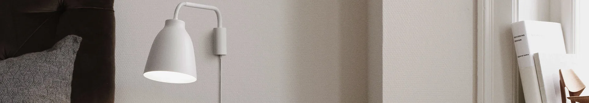 Vit vägglampa som lyser, bredvid en mörkbrun soffa med en grå kudde, samt böcker och en stol i bakgrunden.