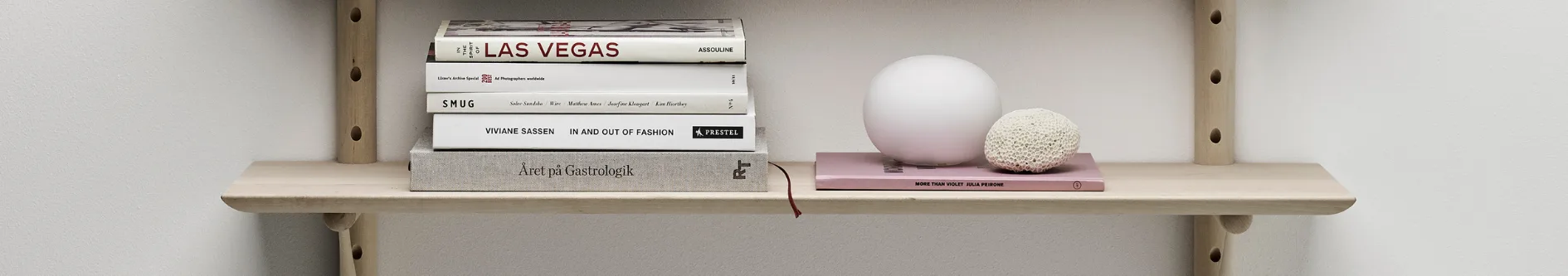 En trähylla med en stapel böcker, en vit sfärisk dekoration och en vit koralliknande föremål på en rosa bok.