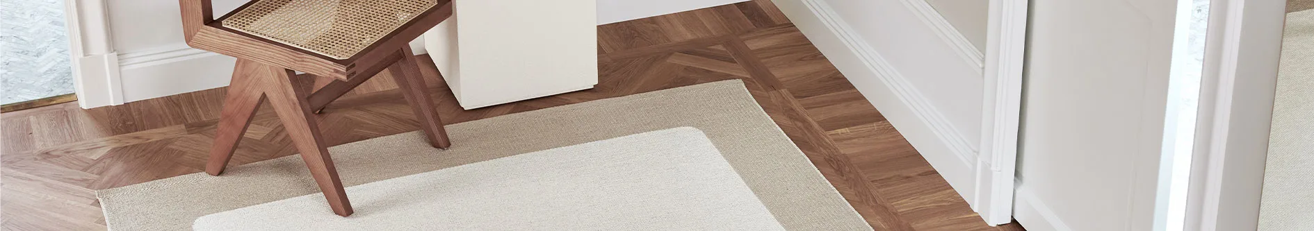 En närbild av ett rum med parkettgolv i trä, en matta i lager (vit över beige) och en träpall med flätad sits.