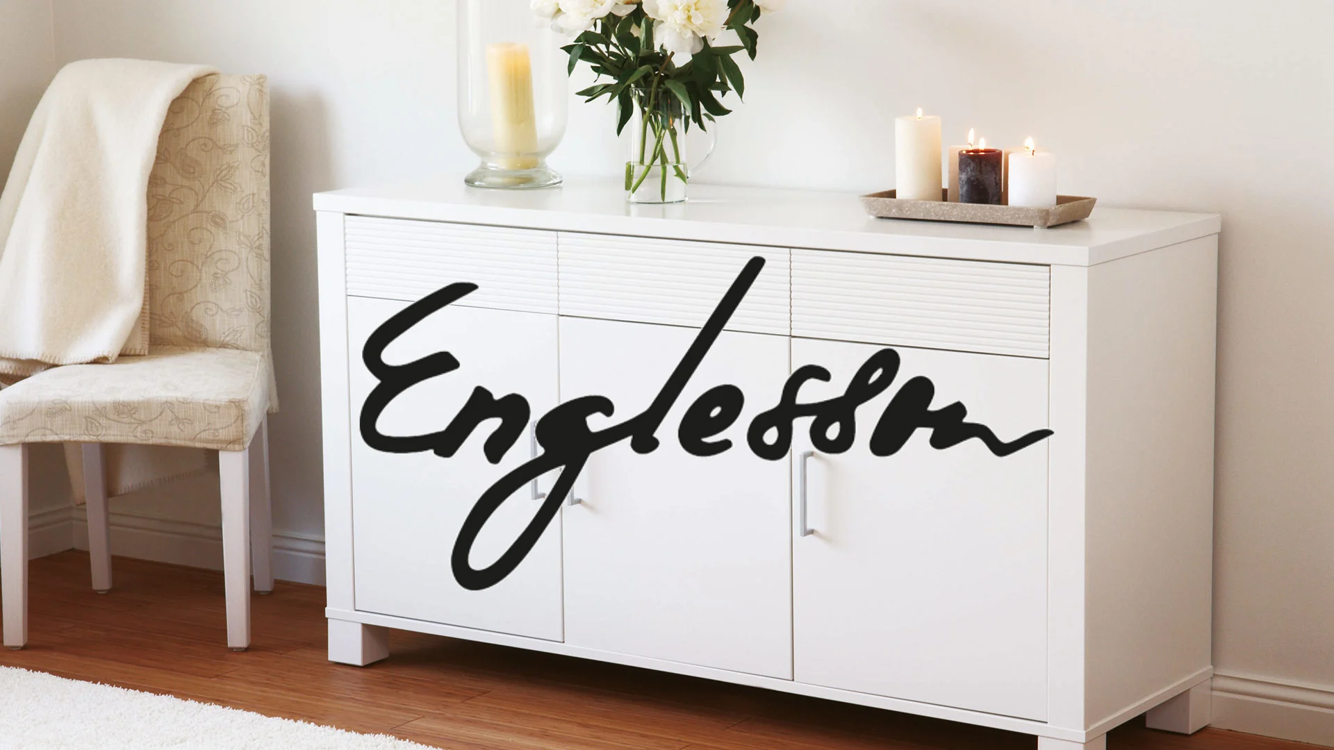 Vit byrå med texten "Englesson" i svart skrivstil, blommor och tända ljus på toppen.