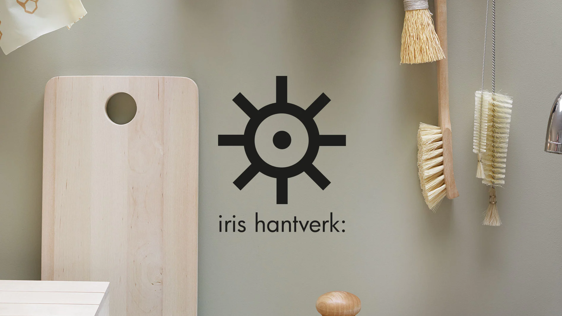Iris hantverk-logotyp på väggen ovanför en skärbräda, kvast och flaskborstar.