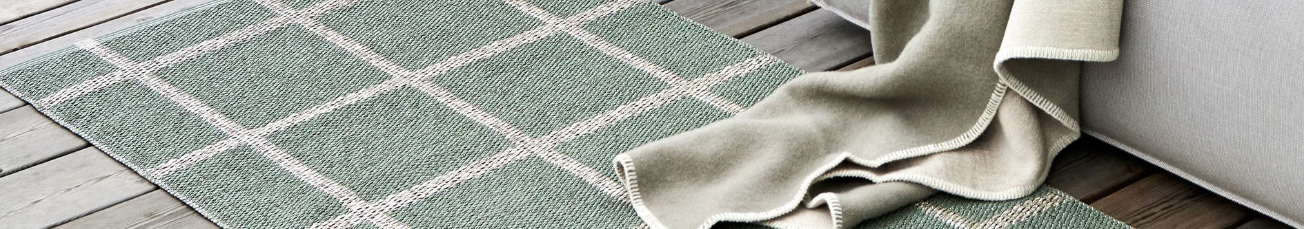 Ljusgrönt vävt rutigt matta på trägolv, med en beige pläd och en ljusgrå soffa delvis synlig.