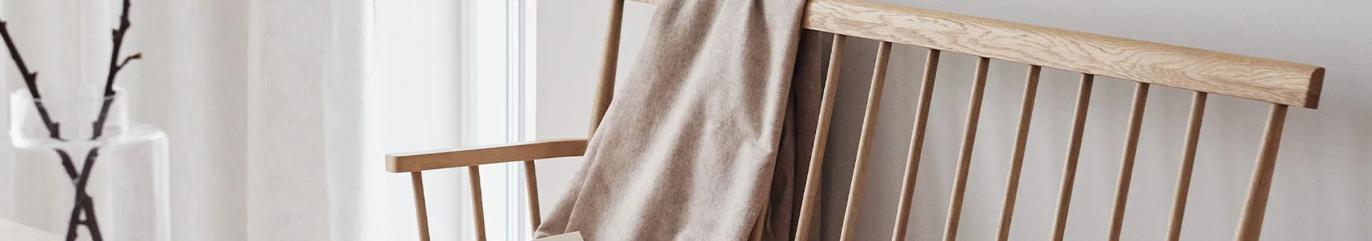 Ljus trästol med en beige pläd draperad över ryggen, med en vas med grenar i bakgrunden.