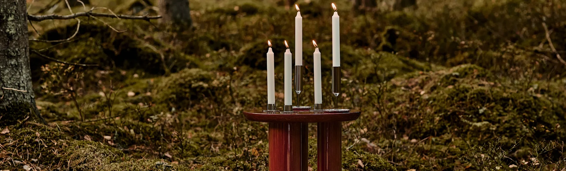 Fem tända, vita ljus i olika höjder på ett rödbrunt bord, mot en mossig skogsbakgrund.