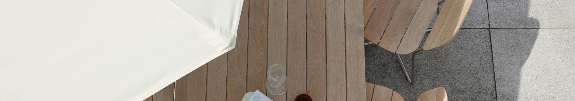 Överbild av ett utomhusbord i trä med två stolar, delvis under ett vitt parasoll, på en stenlagd uteplats. Ett glas och en kopp står på bordet.