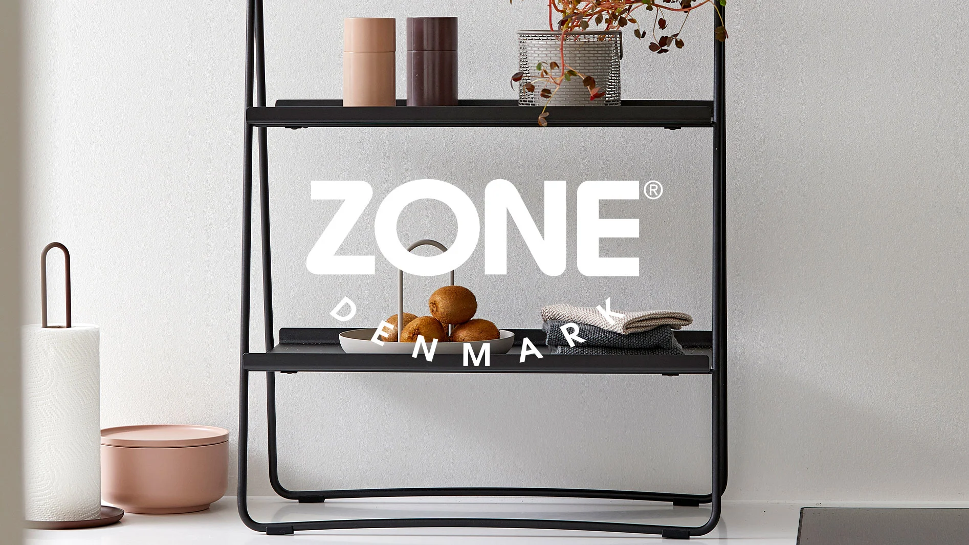 Svart metallhylla med kiwi, handdukar och behållare i ett kök, med Zone Denmark-logotypen överlagrad.