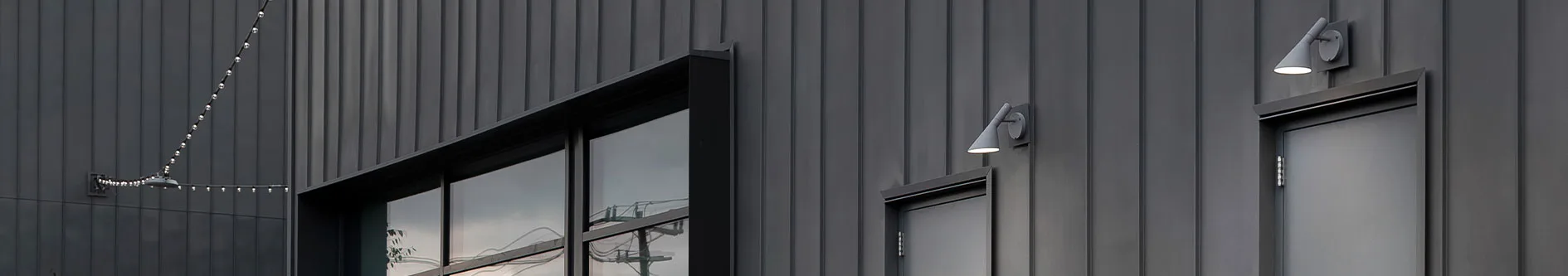 Modern byggnad med mörkgrå panelväggar, upplyst av två vägglampor och en ljusslinga. Har ett stort fönster och en dörr.
