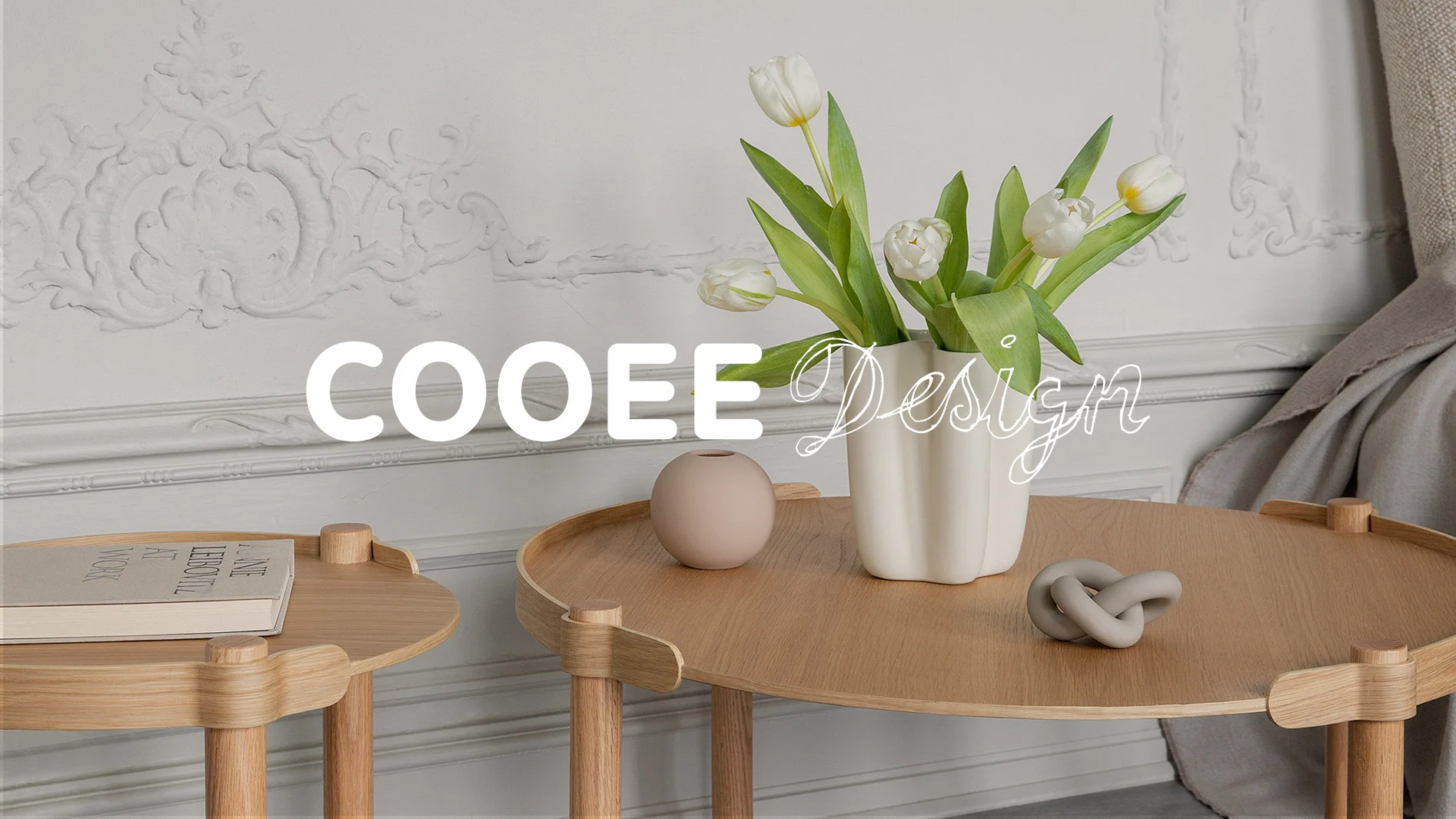 Två runda sidobord i ljust trä med vas med vita tulpaner, en rosa vas, grå dekorativ knut och en bok. Överlagd text: Cooee Design.
