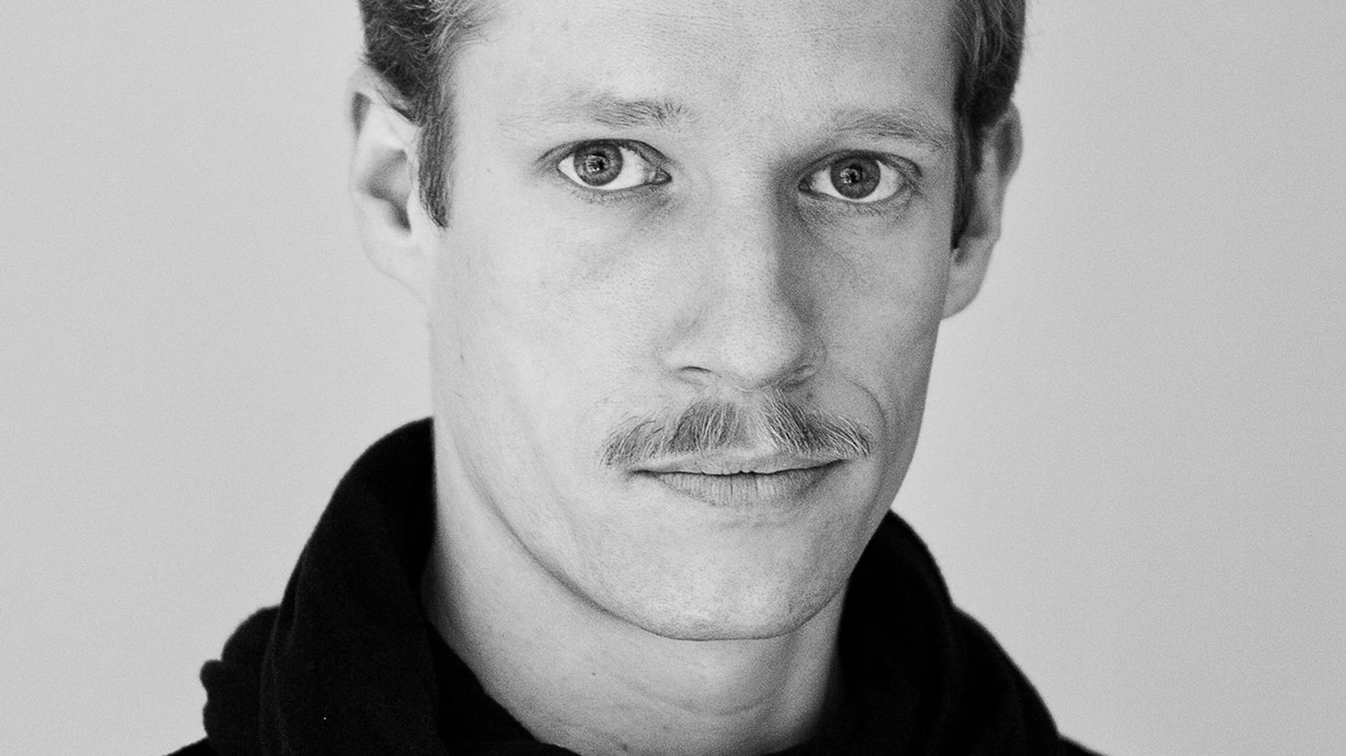 Svartvitt närbildsporträtt av en man med mustasch som tittar rakt på betraktaren.