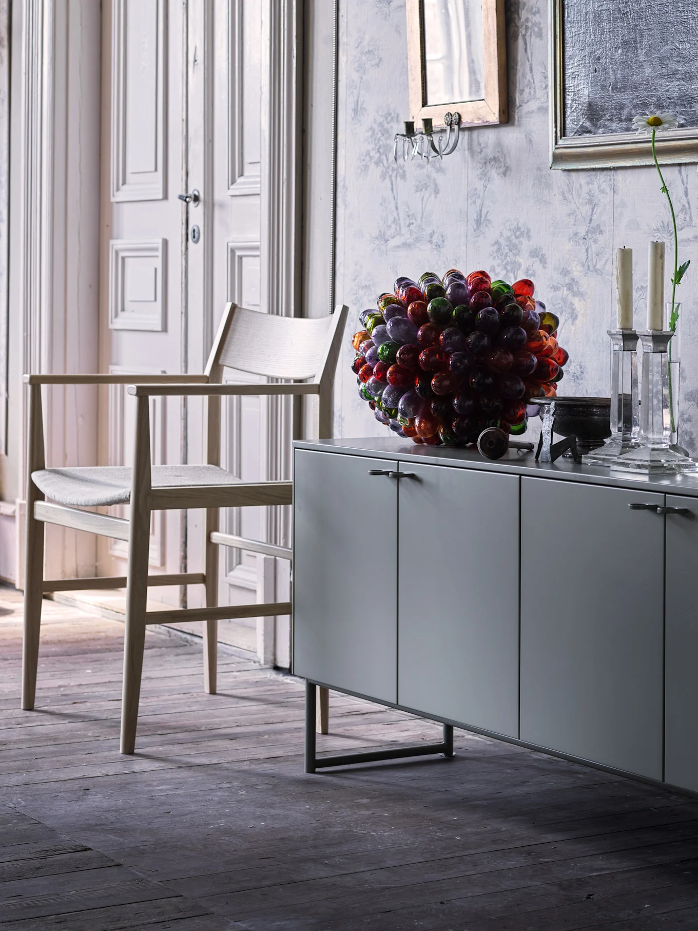 Ett grått sideboard i ett elegant rum, dekorerat med färgrik glasskulptur och vit byst. Vit dörr och mönstrad tapet i bakgrunden.