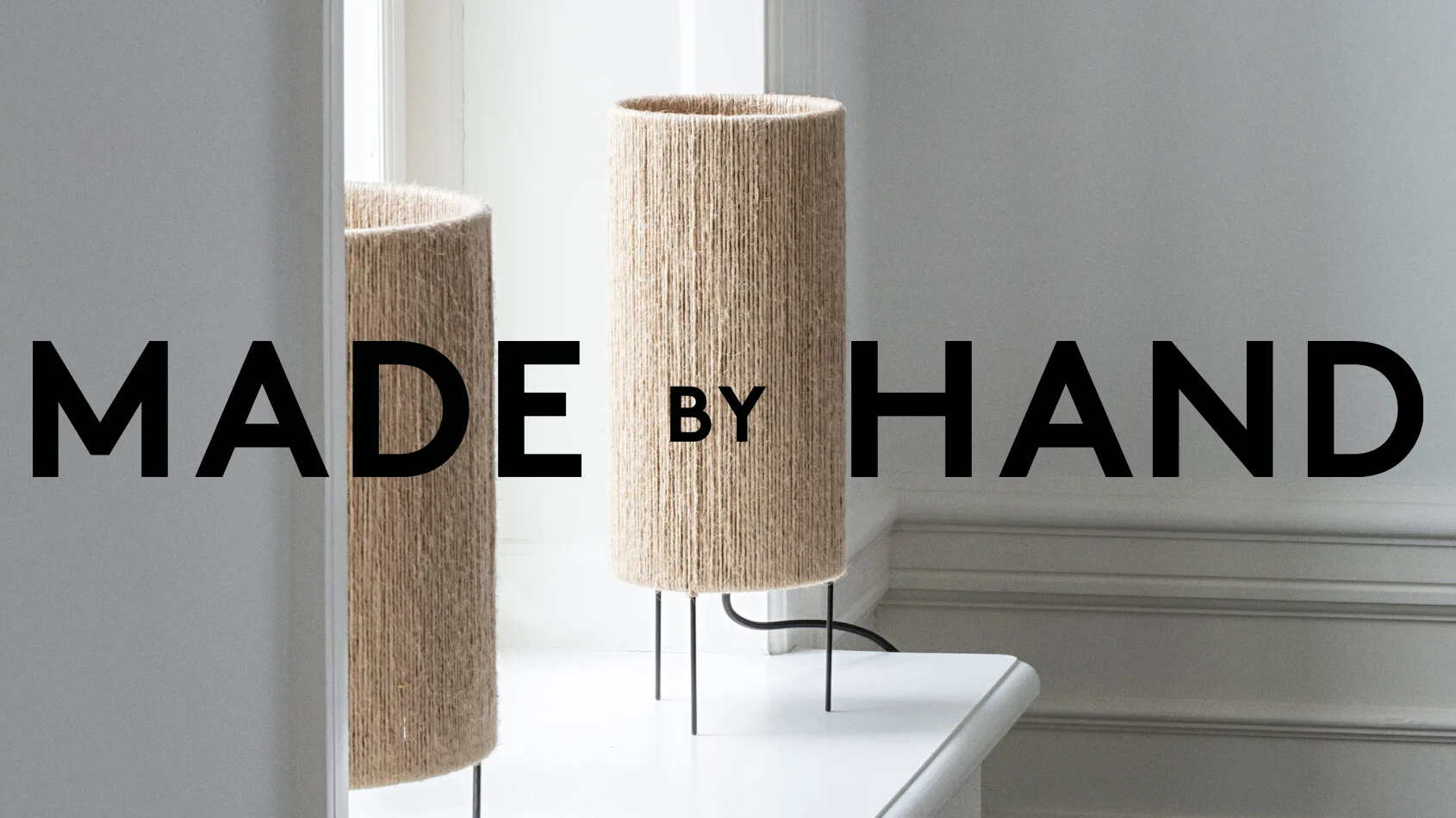 Två cylindriska lampor av naturfiber med svarta metallben. Texten lyder 'MADE BY HAND'.