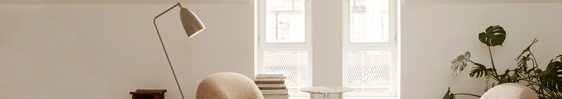 En modern golvlampa, en strukturerad fåtölj, en trave böcker och en monstera-växt i ett ljust rum med fönster.