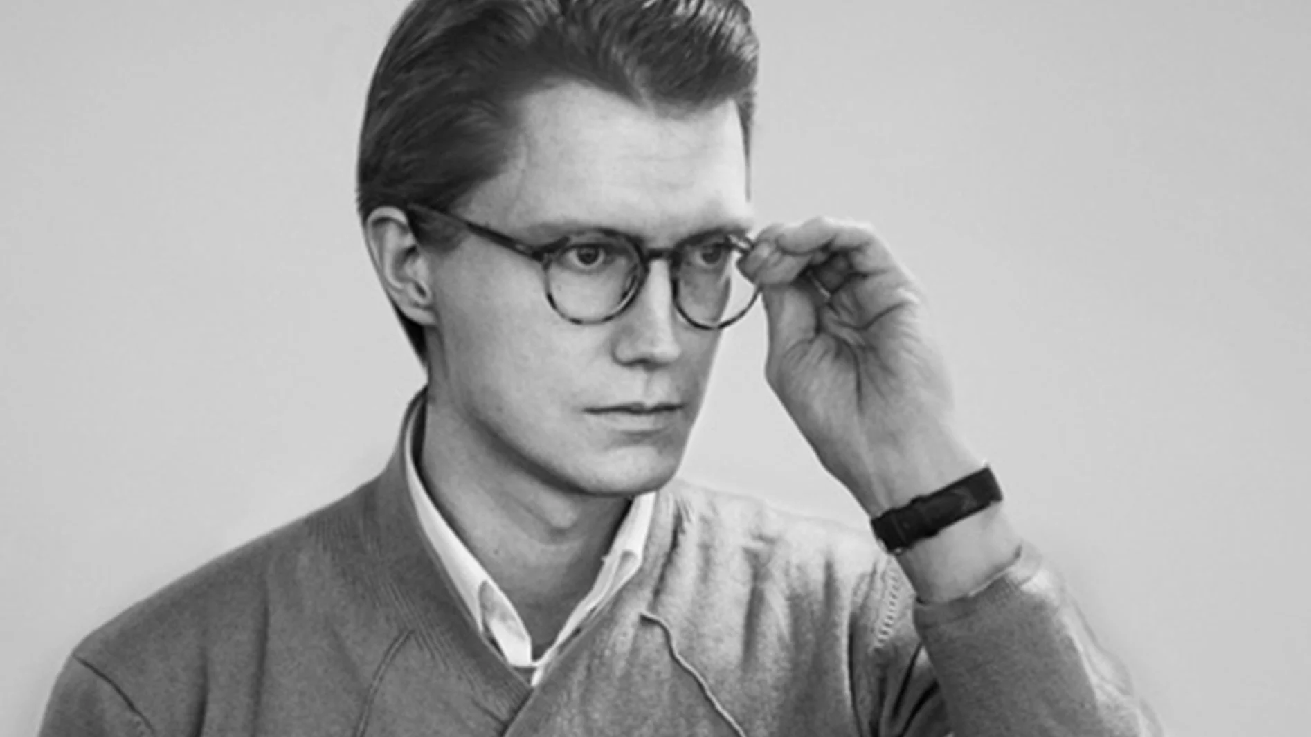 En svartvit bild av en man som justerar sina glasögon och tittar åt höger. Han bär en tröja över en skjorta.